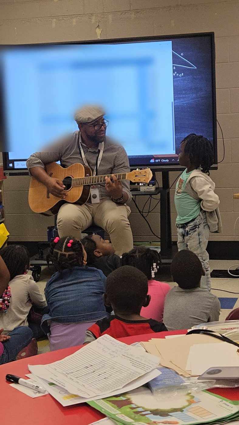 Eli Davis seated with an acoustic guitar, playing for young students gathered on a classroom carpet