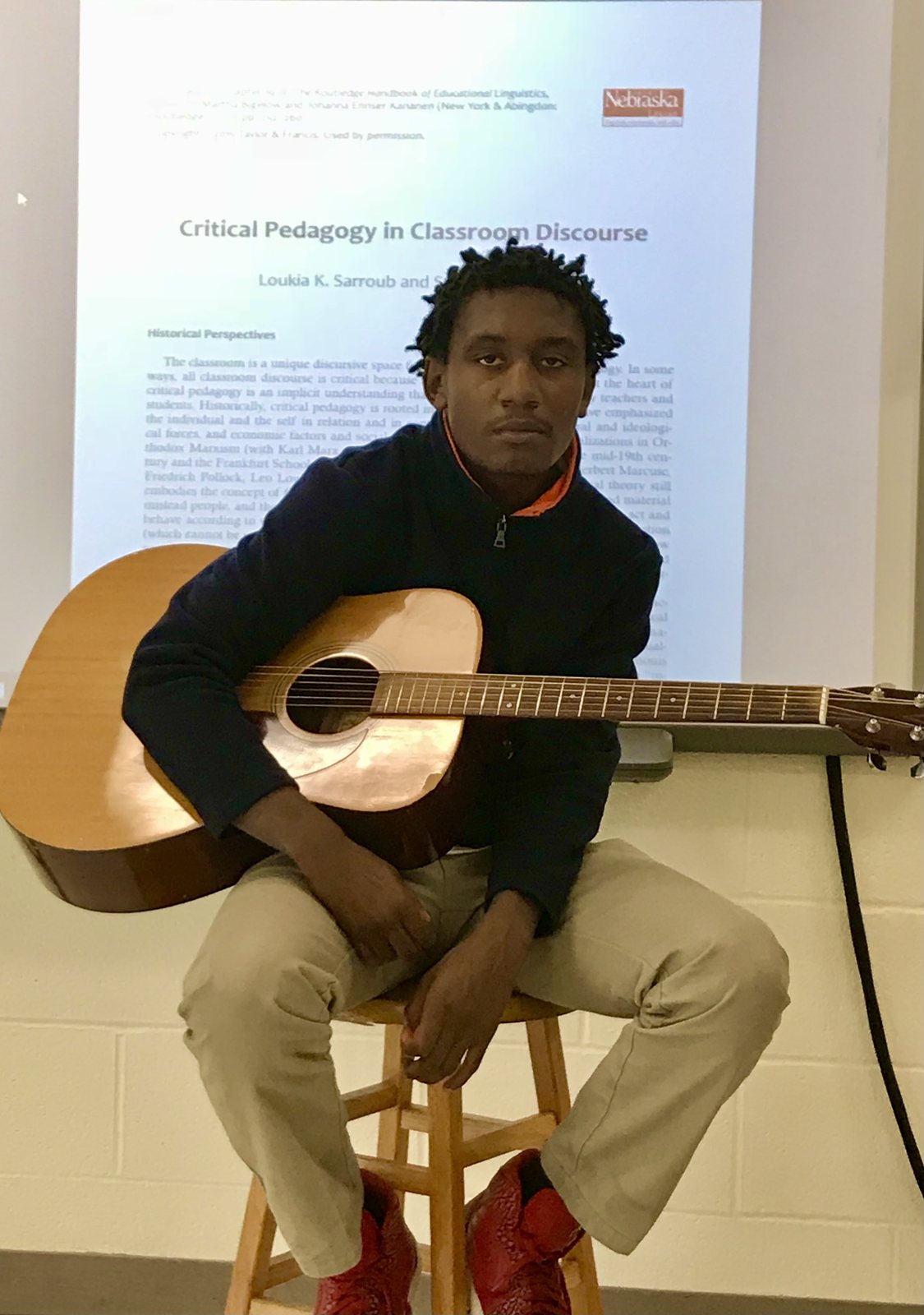 A student seated on a stool holding Eli's acoustic guitar, a projected article on critical pedagogy on the screen behind him