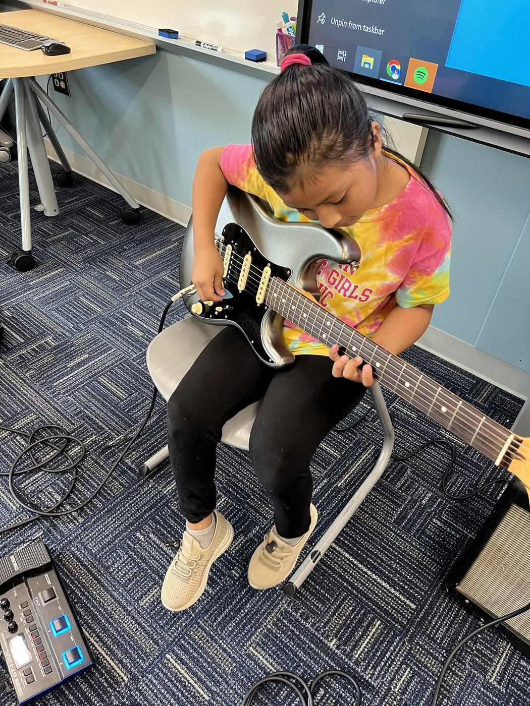 A young student seated in a classroom chair playing a silver electric guitar plugged into an amp