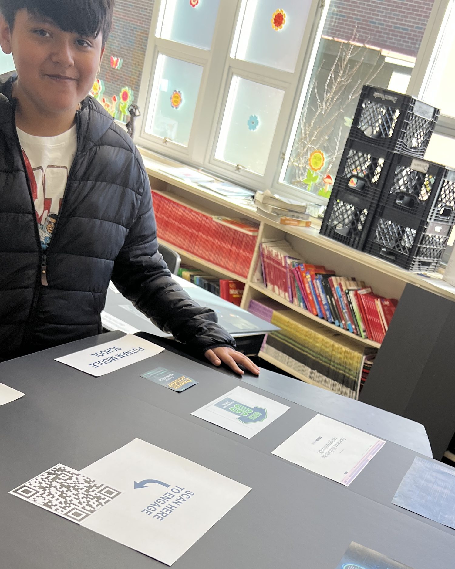 A student stands smiling at his showcase trifold for Striker Academy, hand on the board, beside a scan-here-to-engage sign with its QR code
